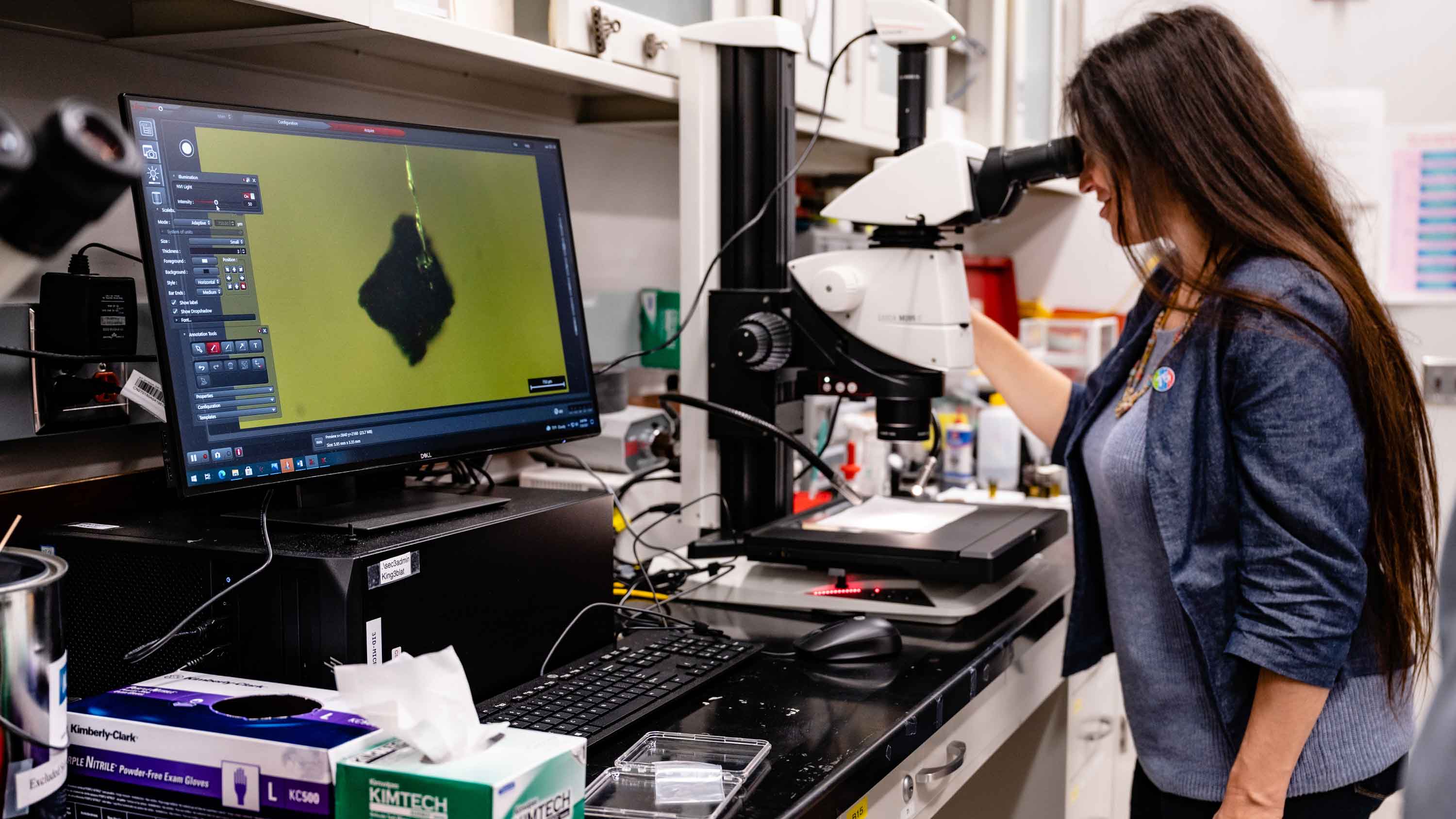 University of Chicago and Argonne beamline scientist Barbara Lavina observes one of the tiny asteroid fragments through a microscope, with the magnified image on the screen beside her. 