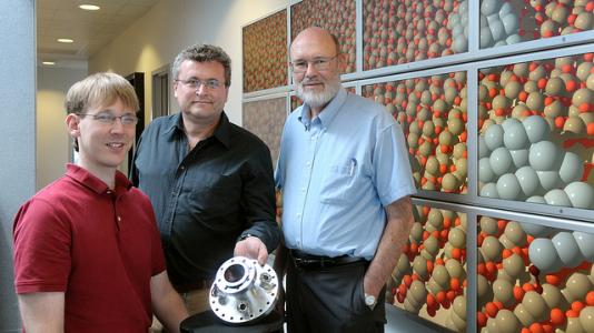 Three Argonne scientists, one of whom is holding a metallic circular instrument, stand in front of a display showing molecular structures.