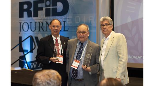 Three men standing aside podium, RFID Journal background. (Image by RFID Journal.)